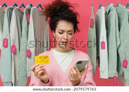 Young sad woman in sweater stand near clothes rack with tag sale in store showroom using mobile cell phone hold credit bank card shopping online order delivery isolated on plain pink background studio