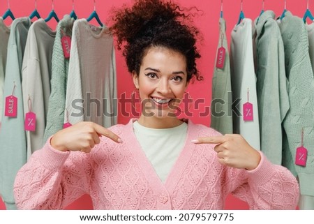 Young smiling happy cheerful female costumer woman wear sweater stand near clothes rack with tag sale in store showroom point index finger on herself isolated on plain pink background studio portrait