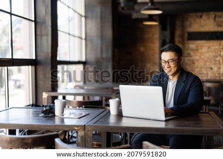 Asian businessman typing on laptop during work in cafe. Concept of remote and freelance work. Smiling adult successful man wearing suit and glasses sitting at wooden desk. Sunny day Royalty-Free Stock Photo #2079591820