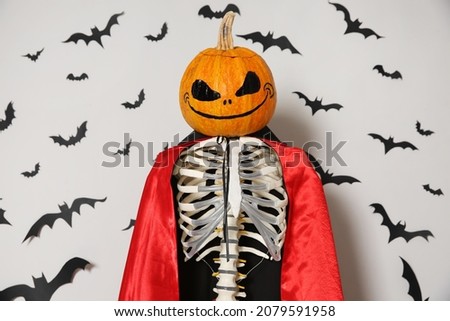 Skeleton in cloak with pumpkin head and paper bats on light wall