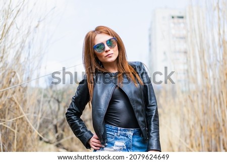 Portrait of a girl with long hair in a leather jacket and sunglasses, in the autumn forest. Young hipster or rocker woman posing on the street. 