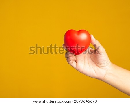 Hand holding of a red heart shape over a yellow background. Space for text. People, healthcare, and love concept