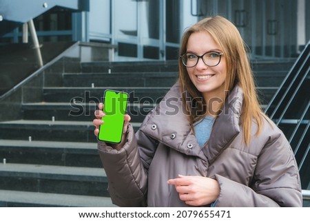 Beautiful smiling caucasian girl with glasses showing blank smart phone green screen