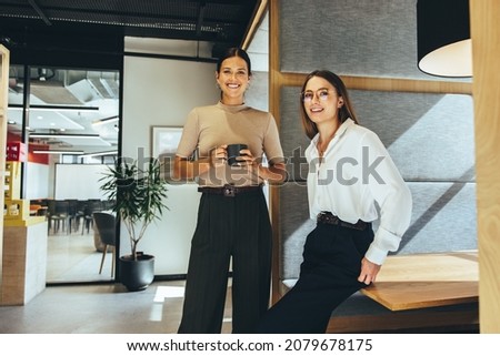 Cheerful female entrepreneurs smiling at the camera while standing in a modern co-working space. Two happy young businesswomen taking a coffee break while working together. Royalty-Free Stock Photo #2079678175