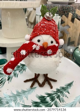 The top down, close up image of a decorative bird wearing a red scarf and hat. The penguin figurine is sitting on a festive plate with green pine trees and next to a green mug with a spoon attached.
