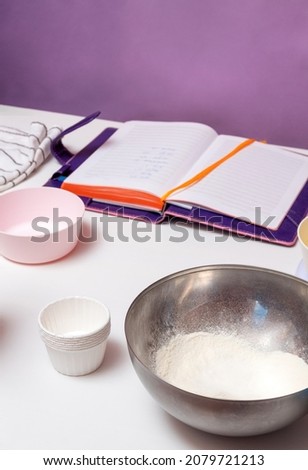 In the foreground is a bowl of flour and cupcake tins. In the background is an open notepad. Selective focus.