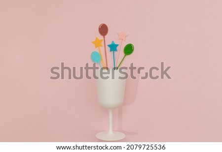 White matte glass with splashes of colourful cocktail cupcake picks on pastel pink background. Minimal Happy birthday flat lay. Celebration card or party concept. Holiday idea.