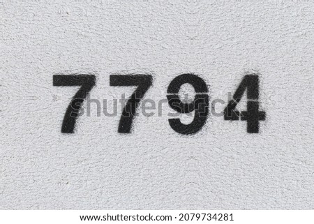 Black Number 7794 on the white wall. Spray paint. Number seven thousand seven hundred ninety four.