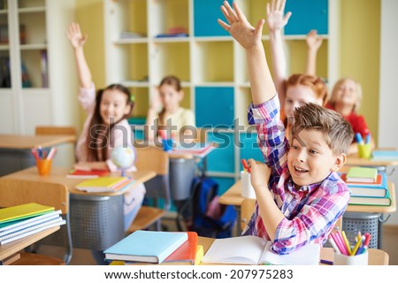 Portrait of cute boy raising hand at workplace with his classmates behind