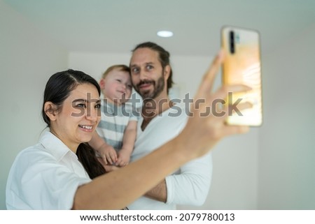 Caucasian happy family smile, look at camera to take picture on phone. Young attractive couple parents, father and mother hold little baby boy child video call and selfie together in bedroom at home.