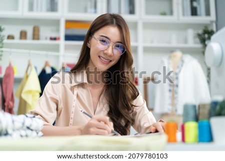 Portrait of Asian tailor woman working on clothes in tailoring atelier. Attractive female fashion designer dressmaker design drawing and sketching out ideas for new fashion collection in workshop room