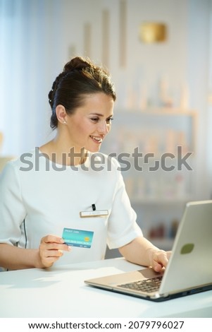 smiling middle aged woman employee with credit card and laptop buying online in modern beauty studio.