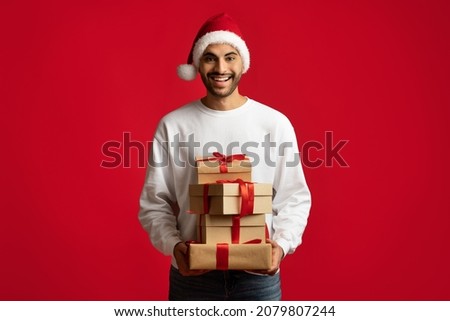 Happy Holidays. Cheerful Arab Man Wearing Santa Hat Holding Stack Of Gifts And Smiling At Camera, Happy Middle Eastern Guy Carrying Present Boxes While Posing Over Red Studio Background, Copy Space