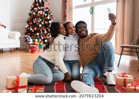 Portrait of cheerful black family of three people celebrating Christmas, taking selfie, smiling excited teenager daughter hugging parents, looking at cellphone camera in decorated living room