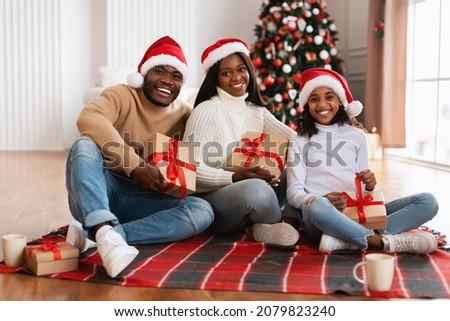 Merry Christmas. Portrait Of Happy Loving Black Family In Red Santa Claus Hat Celebrating Winter Holidays Together, Holding Boxes Sitting On Floor Blanket In Living Room, Posing Looking At Camera