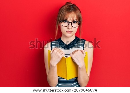 Redhead young woman reading a book wearing glasses relaxed with serious expression on face. simple and natural looking at the camera. 