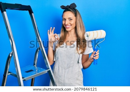 Beautiful hispanic woman by stairs holding roller painter doing ok sign with fingers, smiling friendly gesturing excellent symbol 