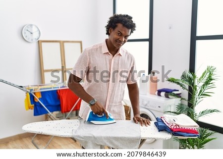 African man with curly hair ironing clothes at home with a happy and cool smile on face. lucky person. 