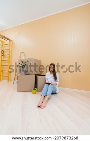 A young woman is sitting on the floor in front of the boxes and picks up a new design for her apartment on a tablet after moving. Housewarming, delivery and cargo transportation.