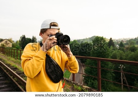 Photographer in stylish casual clothes stands on a bridge in the mountains and takes photos on camera.