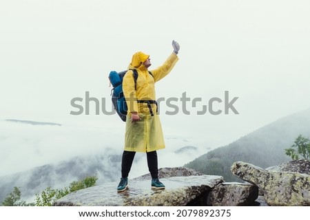 Male hiker on top of a mountain on a rock in rainy weather catches a net on a smartphone on a background of cloudy mountains.