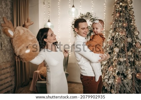 Young family husband, wife and daughter at home near the Christmas tree with gifts. Dad and mom hold the baby in their arms, hugs, joy, happiness. Decorated house, flashlights, quarantine coronavirus.