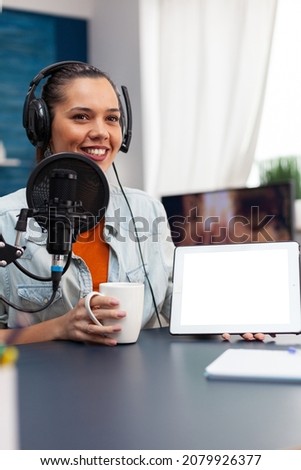 Woman smiling showing digital tablet with white screen. Blogger holding device with mockup template and isolated background while using microphone and headphones to stream podcast.