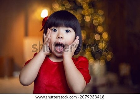 beautiful Korean child girl in a red dress surprised. stands at the Christmas tree on Christmas day