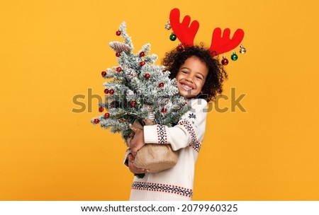 Happy New Year. Little preschool african american kid boy in red deer antlers holding snowy Xmas tree and looking in camera with bright smile, posing against bright yellow studio wall background