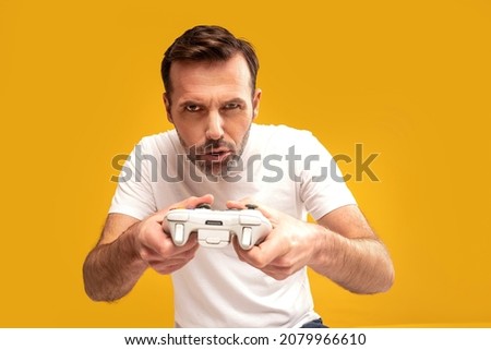 Gambling addiction. Excited mature man holding joystick and playing video games, looking at the camera.  Yellow studio background. A lot of copy space.
