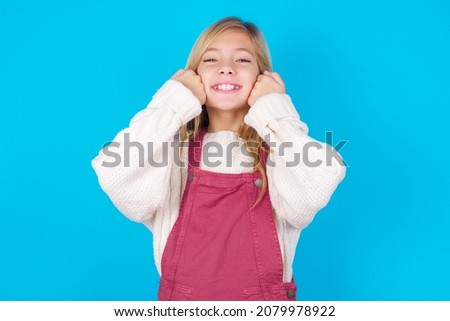 Happy caucasian little kid girl wearing jumpsuit over blue background keeps fists on cheeks smiles broadly and has positive expression being in good mood