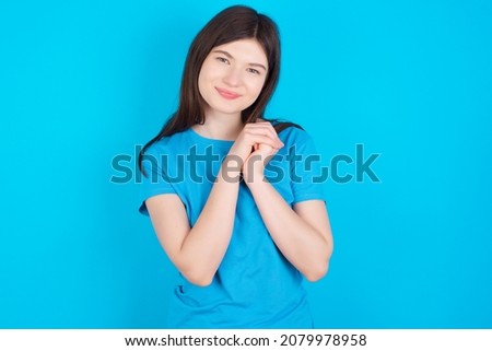 Charming serious Young caucasian girl wearing blue T-shirt isolated over blue background keeps hands near face smiles tenderly at camera