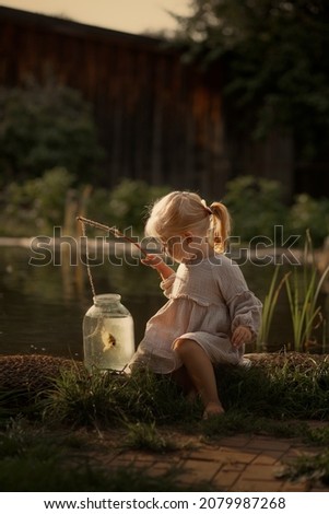 A little girl sits on a pond and catches a fish in a can with a fishing rod