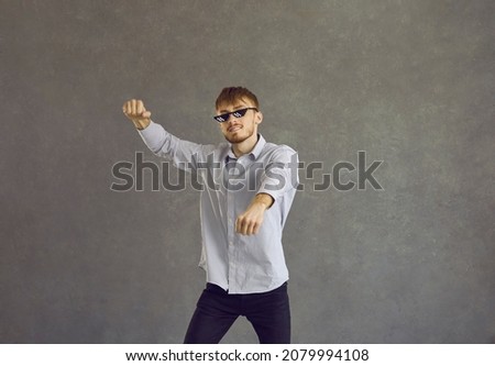 Studio portrait of happy redhead young man in thug life glasses dancing gangnam style. Cheerful guy with red hair wearing funny sunglasses having fun and dancing to music isolated on grey background