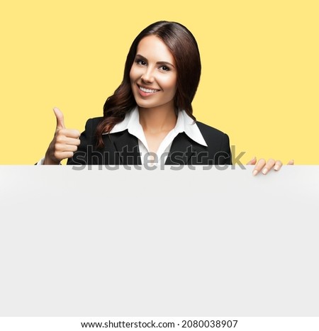 Happy smiling brunette business woman lady in confident black suit, showing thumb up hand gesture, standing behind, peeping from blank banner or mock up ad signboard, over yellow color background.