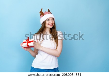 Pretty young woman in christmas deer antlers and white t-shirt isolated on a blue background, holding a gift box for christmas and new year, smiling with white teeth with happiness.