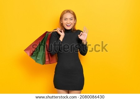 Smiling young Asian woman holding shopping bags and gesturing okay sign isolated over yellow background