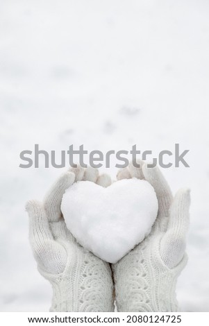 A snow heart in the hands wearing knitted gloves against the background of a snow cover. Royalty-Free Stock Photo #2080124725