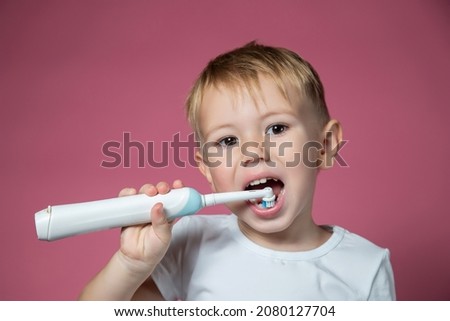 Smiling caucasian little boy cleaning his teeth with electric sonic toothbrush on pink background.