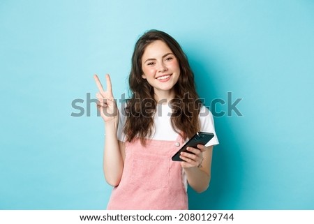 Online shopping. Cute and cheerful young woman smiling, holding smartphone and showing peace v-sign at camera, standing against blue background