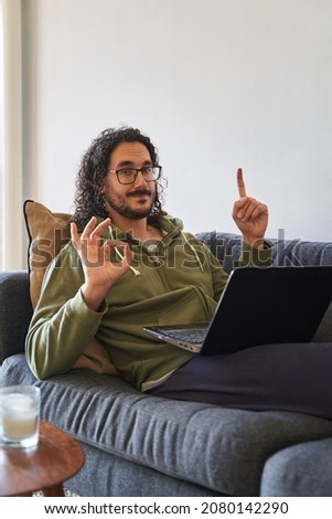Vertical medium shot of young man on the sofa with laptop smiles and points away and makes ok sign