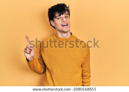 Handsome hipster young man wearing casual yellow sweater with a big smile on face, pointing with hand finger to the side looking at the camera. 