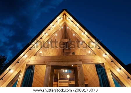 Roof with garlands lights of a small wooden lodge house with glass door and window at night time. The concept of glamping and idyllic holidays Royalty-Free Stock Photo #2080173073