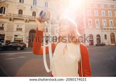Happy young woman traveler photographer holds camera on European street and takes picture. Summer sun light.
