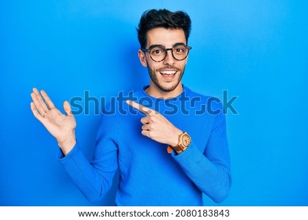 Young hispanic man wearing casual clothes and glasses amazed and smiling to the camera while presenting with hand and pointing with finger. 