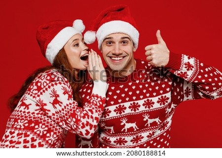 Young couple friends two man woman in sweater hat whispering gossip and tell secret behind her hand share news show thumb up isolated on plain red background. Happy New Year 2022 celebration concept