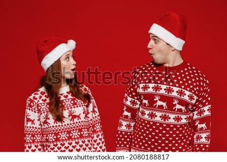 Young shocked amazed couple friends two man woman 20s in sweater hat look to each other isolated on plain red background studio portrait. Happy New Year 2022 celebration merry ho x-mas holiday concept