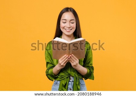 Smiling cheerful attractive beautiful young brunette asian woman 20s wearing basic green shirt standing reading holding in hands book isolated on bright yellow colour background, studio portrait