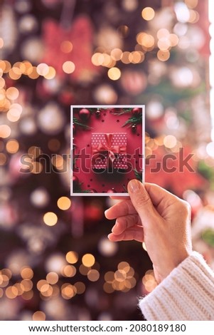 Female hand holding merry Christmas card vintage film photo holiday greeting photograph picture with red xmas present gift on decorated blurred New Year tree lights background. Vertical