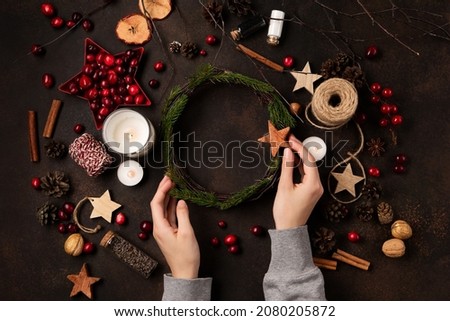 Women's hands make natural Christmas wreath. Yule winter solstice concept. Tree branches, red berries, candles on wooden background, top view, copy space. Zero waste Christmas, plastic free holidays.